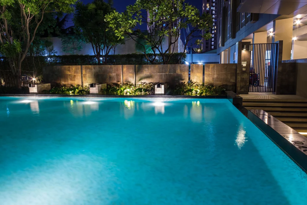 Piscina iluminada à noite com refletores subaquáticos, destacando o brilho da água e o paisagismo moderno ao redor.