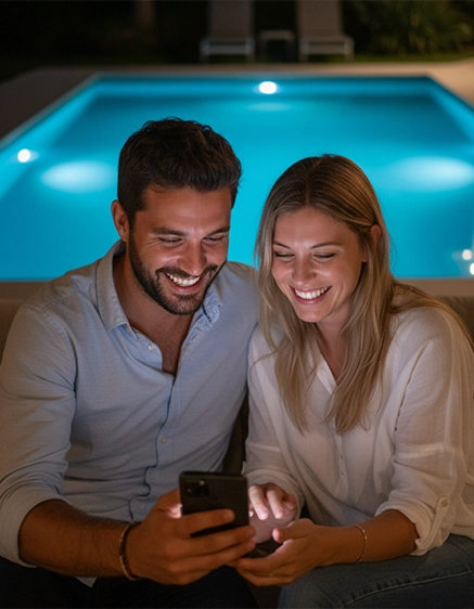 Casal sorridente utilizando o aplicativo à noite em área de lazer com piscina iluminada ao fundo.
