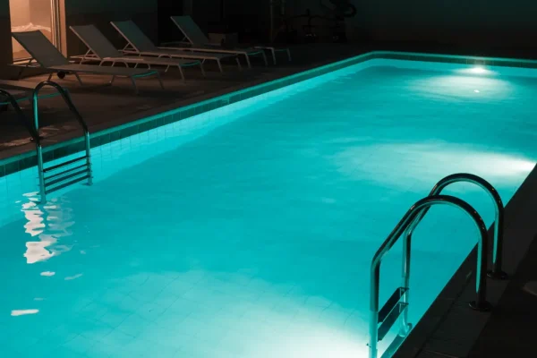 Nighttime swimming pool illuminated with underwater lights, featuring turquoise water, stainless steel ladders, and a calm residential outdoor setting.