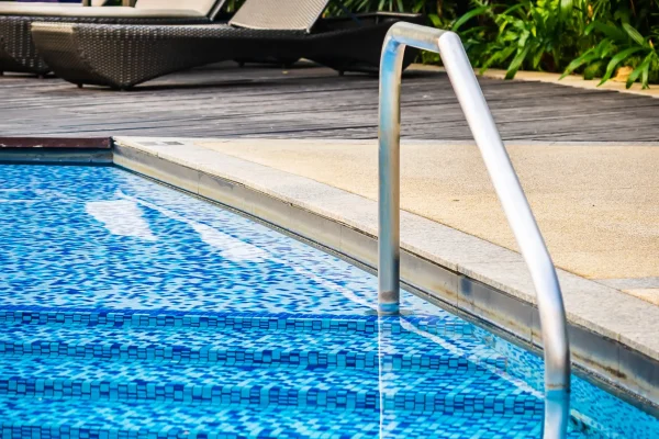 Piscina ao ar livre com escada de aço inox, degraus revestidos em pastilhas azuis e espreguiçadeiras em ambiente com paisagismo tropical.