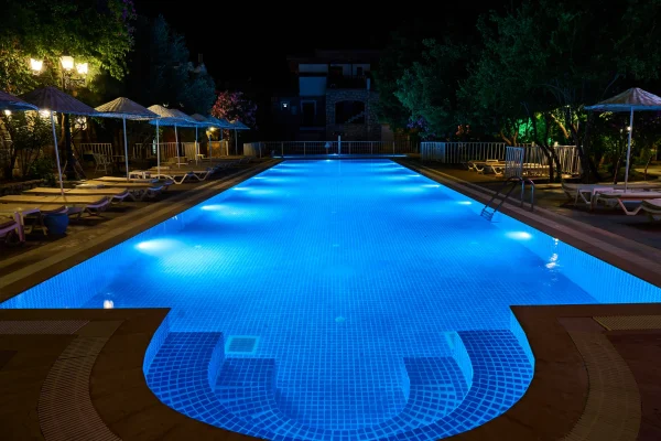 Piscina de hotel iluminada à noite com luzes azuis subaquáticas, espreguiçadeiras e guarda-sóis ao redor, criando um ambiente elegante e acolhedor.